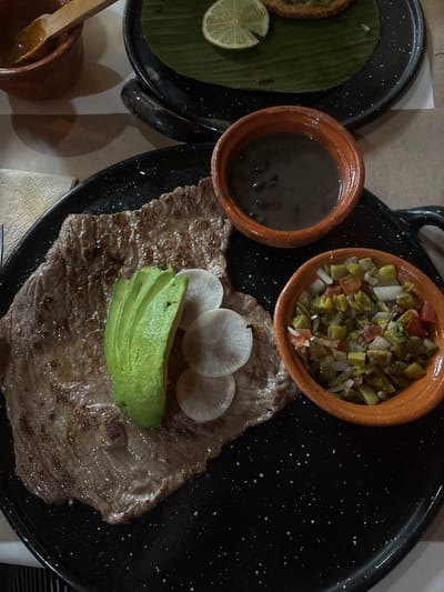 A close-up, overhead view captures a dining table set with Mexican cuisine in San Sebastián Xolalpa, Mexico. The central subject is a large, oval black plate with white speckles, holding a grilled, thinly cut piece of meat, likely flank steak, displaying char marks. This meat is topped with a fanned arrangement of four green avocado slices and three thin, white, circular radish slices. On the same plate, two small, round, terracotta-colored bowls are present: one contains black beans, and the other holds a diced salad composed of green nopales or chiles, red tomatoes, and white onions. In the background, partially visible, is another black, speckled plate with a green banana leaf and a single lime wedge. A round, griddle-cooked item is partially visible on this banana leaf. To the upper left, a small, reddish-brown clay pot with a wooden spoon contains a reddish sauce. The table surface is a light brown.