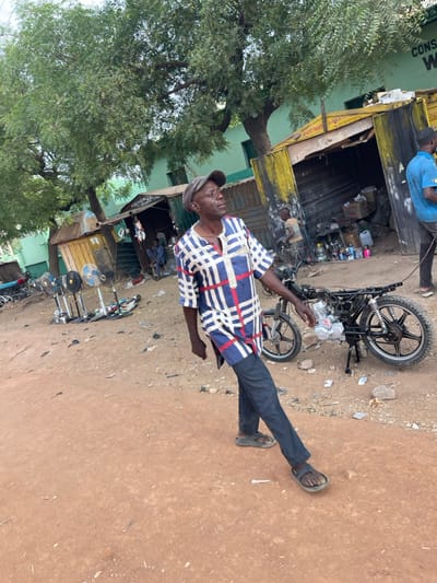 An older man in a checkered shirt, cap, and dark pants stands next to a partially disassembled motorcycle. He is on a dirt ground near a building with a yellow facade. Another person is seen working on something in the background. Trees are visible in the foreground and to the side. Several fans are on stands nearby.