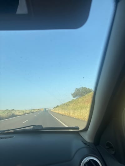 The image is taken from inside a car, looking through the windshield. The car is on a highway, with a grassy verge and occasional trees to the side. The sky is blue with some clouds. The interior of the car is visible on the sides of the frame, including an air vent. The exact location within the city of Sidi Slimane Moul Al Kifane, Morocco is not discernible from the image. There are no visible people.