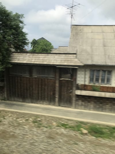 This image captures a scene in Vicovu de Sus, Romania, characterized by significant horizontal motion blur consistent with being photographed from a moving vehicle. The environment consists of several buildings, trees, and ground features under an overcast sky.

The upper portion of the frame reveals an overcast sky with a diffused, light grey tone and visible cloud formations. A prominent metallic Yagi antenna, consisting of multiple horizontal elements, extends vertically from the roof of the rightmost visible building.

The building on the right features a light grey pitched roof, appearing to be constructed from corrugated sheets, showing signs of weathering and dark staining. The upper facade of this building is light-colored, possibly white or cream, with visible vertical paneling. A multi-pane window with a light-colored frame is present in this section. Below the window, a dark brown, horizontally oriented wooden planter box or decorative element is affixed. The lower section of this building's wall is clad with vertical dark wooden planks.

Adjacent to the left of this structure, a building with a dark wooden facade is visible. It possesses a light-colored pitched roof, similar in material and condition to the adjacent structure. The primary feature of this facade is a large section composed of vertically oriented dark wooden planks, which appears to be a gate or garage doors. Above this wooden section, a series of horizontal, slatted openings or small windows are observable.

Further to the left, a substantial tree with dense green foliage dominates the frame, exhibiting pronounced motion blur. A smaller tree or dense bush is situated behind the central wooden structure, positioned to the left of the antenna.

The foreground of the image consists of a dark grey paved surface, likely a road, bordered by a lighter-toned strip on its immediate lower edge. Below this paved strip, the ground transitions into an area composed of gravel, dirt, and sparse patches of green vegetation, all rendered with motion blur. No individuals, animals, or other vehicles are present in the visible scene.