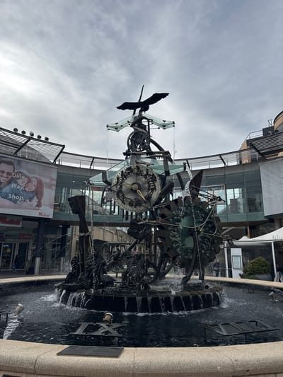 The image shows a complex, multi-tiered kinetic sculpture fountain in the center of a circular pool, located in Hornsby, Australia. The sculpture is made of dark metal and features gears, figures, and what appears to be a large clock face. Water cascades from various levels of the sculpture into the pool below, creating a dynamic display. Surrounding the fountain is a stone-edged pool with some metal elements submerged in the water, including Roman numerals. In the background, there is a modern building with glass facades, a large billboard advertising "Westfield" with images of a couple, and shops like "Coles" and "Kmart." The sky is overcast, and the overall lighting suggests it is daytime. Several people are visible in the background, some walking near the shops and others near an outdoor seating area. The environment appears to be an urban public space, likely a shopping center or plaza.