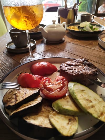 A close-up shot shows a plate of food on a wooden table. The plate contains a grilled steak, sliced tomatoes, grilled eggplant, and zucchini. To the left, a glass of amber-colored liquid with ice and condensation is visible, along with a small grey cup and saucer. In the background, a white teapot, a salad bowl, and cutlery in a holder are on the table. A blurred figure of a person is visible in the far background. The scene is indoors, likely a restaurant, with natural light illuminating the setting. The Uglich State Historical Architectural and Art Museum is a possible location where such a meal might be served, given the historical context of Uglich, Russia.