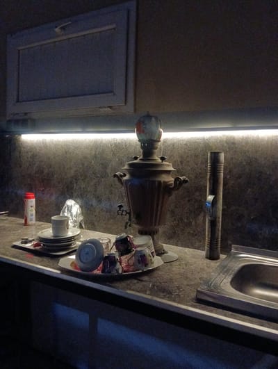 The image depicts a dimly lit kitchen scene with a focus on a traditional samovar and tea set. The samovar, a large, ornate metal vessel used for heating water for tea, is centrally positioned on a countertop. Several teacups and saucers are arranged around it, some stacked and others individually placed. The teacups appear to be decorated with floral patterns. To the left of the samovar, a red and white cylindrical container, possibly for sugar or tea leaves, is visible. Behind the samovar and tea set, a textured, dark-colored wall is illuminated by a strip of white LED lighting running horizontally above the countertop. To the right of the samovar, a tall, cylindrical metallic object stands upright, its surface showing ribbed details. Further to the right, a stainless steel sink is integrated into the countertop. Above the countertop, a closed cabinet with a light-colored front and a subtle handle is present. The overall environment suggests a functional kitchen space, possibly in a residential setting in Türkan, Azerbaijan, given the prompt. The lighting creates a somewhat atmospheric and shadowed ambiance. There are no people visible in the image.