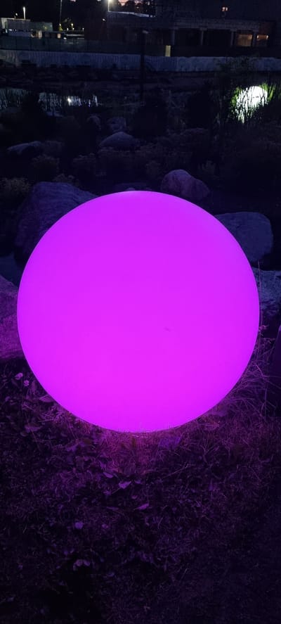 The image shows a large, glowing pink orb situated in an outdoor setting at night. The orb is the dominant subject, emitting a vibrant magenta light. It appears to be a decorative garden light.

The background is dark and somewhat indistinct, suggesting it's nighttime. To the left of the orb, there's a glimpse of a building with windows that are illuminated, indicating interior lighting. There are also some large rocks or stones scattered around, and dense, dark foliage or grass encroaching on the scene, particularly on the right side. The overall impression is one of a garden or landscaped area at night, possibly a yard or patio.

There are no people visible in the image. The scene is static, with the orb serving as a point of light in the darkness. The time of day is clearly night due to the darkness and the artificial lighting from the orb and the building. The weather is not discernible from the image. The location cues from the building and the general aesthetic hint at a residential or possibly commercial establishment with outdoor landscaping. No text is visible in the image.