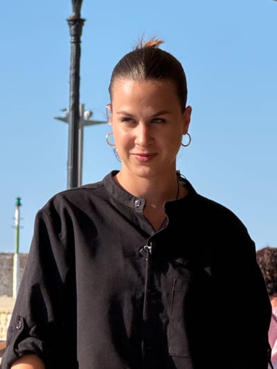 A medium close-up shot shows a young woman with dark hair pulled back in a bun, facing slightly to her left. She is wearing a black, collared shirt with buttons at the neck and cuffs. She has hoop earrings and is looking downwards and to her left with a subtle smile. The background is a bright blue sky with the upper portion of a black lamppost visible on the left side of the frame. The image appears to have been taken outdoors in Chania, Greece.