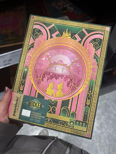 The image shows a "Wicked" unlined hardcover journal, a product of Paperblanks, being held in someone's hand. The journal's cover design features a pink and gold patterned circle, with elements that seem to reference the musical "Wicked". It includes silhouettes of the main characters and a castle. The surrounding design incorporates pink and green geometric patterns with gold detailing. A price tag of 27.95€ is visible in the background, suggesting the journal is being sold in a retail environment. The hand holding the journal has a glittery nail polish. Based on the price in euros, it is probably in Europe. Based on the price tag that mentions the price is "27.95€", it indicates the picture was probably taken in Cordoba, Spain.