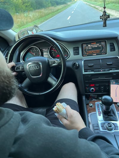 The image depicts the interior of a car, likely an Audi Q7 based on the steering wheel and dashboard design, as it travels down a paved road. The road is bordered by green foliage, suggesting a rural or semi-rural setting. A male driver, with a beard visible, is holding the steering wheel with his left hand and a sandwich with his right. The sandwich appears to be a wrap with ham and cheese.

The car's dashboard is illuminated, showing the current speed at 84 km/h and the outside temperature at +10.5°C. The infotainment system displays "музика люмень 2025" (music lumen 2025) and song titles in Cyrillic. A car accessory, possibly a rosary or pendant, hangs from the rearview mirror. A camouflage patterned pouch with a red and white emblem is visible on the dashboard.

The central console features the gear selector, climate control, and navigation buttons. A smartphone is mounted on the console, displaying a navigation interface with the destination "Тернопіль" (Ternopil) and a distance of 25 km. The driver is wearing a dark hoodie and black shorts. The overall scene suggests a journey is in progress. The location mentioned, Darakhiv, Ukraine, aligns with the presence of Cyrillic text on the infotainment system and the navigation destination.
