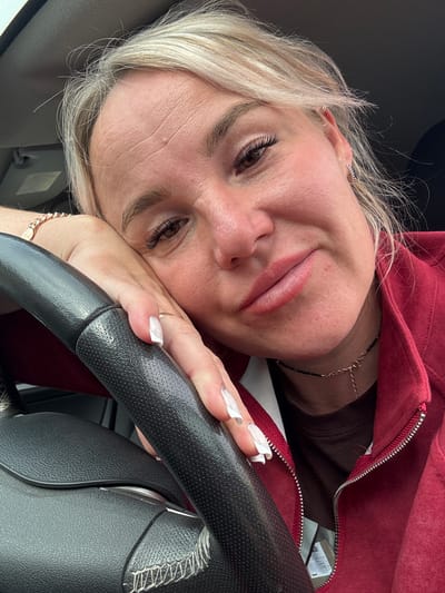 A close-up, waist-up shot features a woman with blonde hair, resting her head on her left hand, which is placed on a black steering wheel. She is looking towards the camera with a slight smile. The woman is wearing a dark brown shirt and a red zip-up jacket. Her left hand displays long, white acrylic nails. A delicate gold necklace is visible around her neck. The background is blurred and appears to be the interior of a car. The scene is likely set within the city of Chastyye, Russia, as indicated by the provided information.