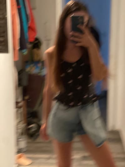 A blurred image shows a person holding a phone, apparently taking a selfie in a mirror. The person is wearing a black top with white patterns and blue denim shorts. The background is indistinct, with hints of clothing and other objects on a shelf or table. It is not possible to determine the location in the city of Paiporta, Spain from the image.