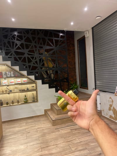 An indoor view shows a staircase with display cabinets built into the steps. A decorative black metal screen adorns the side of the stairs. The staircase leads to an upper level, indicated by the presence of a window and plants. A partially visible hand holds a gold vape device. The flooring is light wood-grained. The room has a modern design with white walls and a corrugated metal ceiling.