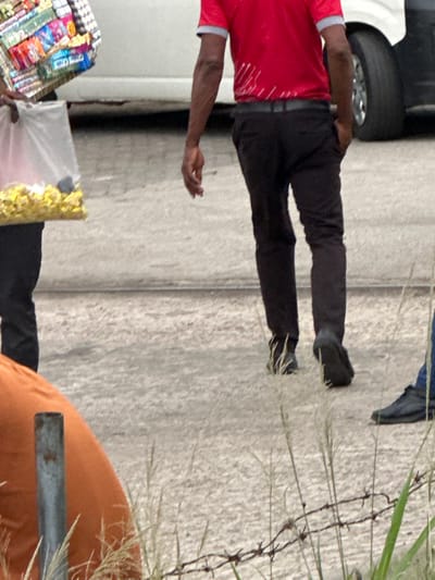 A man stands on a concrete surface beside a car. He wears a red shirt, black pants, and dark shoes. In his hands, he carries a tray of snacks and a bag filled with yellow items. Nearby, an orange object rests on the ground. A barbed wire fence and tall grass are visible, adding depth to the scene.