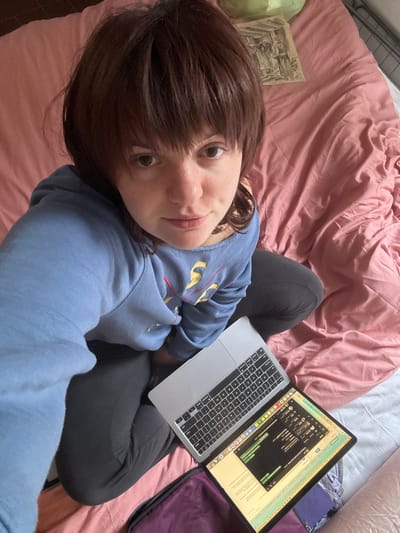 A high-angle selfie depicts a single female individual seated cross-legged on a bed indoors. The individual has medium-length dark brown hair with bangs, light skin, and a neutral expression while looking directly at the camera. She wears a light blue long-sleeved sweatshirt with gold and red embroidery on the chest, and dark trousers. A silver Apple MacBook Pro laptop, open and displaying multiple windows including a chat application and text, rests on the bed between her legs. The bed is covered by a rumpled pink duvet, with a white sheet visible underneath on the right side. A small piece of paper with a black and white drawing of multiple figures is positioned on the pink bedding above and to the right of the individual's head. A purple fabric case is partially visible beneath the laptop. A light green crinkled object and a metallic railing are marginally visible in the upper right background. The image provides no visual information to determine a specific location within Cortona, Italy.