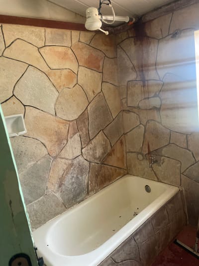 This image depicts the interior of a bathroom within the city of Pirenópolis, Brazil, focusing on a bathtub and surrounding walls. The scene is presented from a slightly elevated perspective, looking into a corner.
The central subject is a white, rectangular bathtub positioned against two walls, occupying the lower half of the frame. The bathtub's interior appears slightly soiled with dark specks and debris. Its drain is visible at the far end.
The walls surrounding the bathtub are clad with irregular, multi-colored stone or stone-patterned tiles, primarily in shades of beige, brown, and grey, with dark grout lines. An electric shower head, identified by the visible "DUCHA FAME" branding, is mounted high on the left wall, near the ceiling. It is white and connected by a hose assembly. Directly below the shower head, a white, plastic or ceramic soap dish is affixed to the wall. On the right wall, above the far end of the bathtub, a single silver-colored metal faucet is installed.
Above the faucet, and extending from the ceiling line downwards along the right wall and into the corner, are prominent dark reddish-brown stains, suggesting water damage or rust. Similar, less pronounced staining is visible along the top edge of the left wall. The ceiling appears to be paneled, possibly wood or PVC, with a dark trim piece where it meets the walls.
Bright natural light enters from the right side of the frame, possibly from a window, casting horizontal shadows across the right wall. A section of a light green door frame or partition is visible on the far left edge of the image, partially obscuring the view of the wall and bathtub. The floor, visible on the far right, appears to be a reddish-brown color, potentially stained concrete or tile. No people are present, and no actions or interactions are depicted.