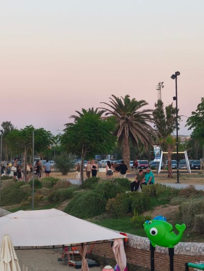 The image depicts a scene in the city of San Salvo, Italy, likely during the late afternoon or early evening, given the soft, pastel colors of the sky. In the foreground, a white canopy tent covers a sandy area, with a beige umbrella to its left. A child's playground element shaped like a green fish is visible on the right, mounted on a brick wall.
In the middle ground, a grassy slope is covered with various types of vegetation. Several people are gathered on this slope, some standing and others sitting. A couple, a man in a turquoise shirt and a woman in a dark green top, are seated together. Further back, a group of people are standing and conversing.
The background reveals a parking area filled with cars. Beyond the parking lot, there are trees, including palm trees, and buildings. Two tall light poles with multiple lamps are also prominent features in the scene, suggesting a public space like a park or a waterfront promenade. The overall atmosphere appears relaxed and social.