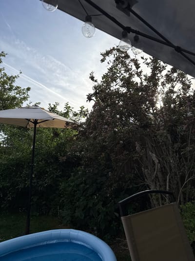 The image, apparently taken outdoors in Hazlemere, United Kingdom, shows a garden scene with a partially visible inflatable blue swimming pool in the lower right corner, positioned near dense green foliage. An outdoor chair with a beige fabric back is adjacent to the pool. Two separate patio umbrellas are present. One, with string lights attached, is mostly out of frame in the upper left. The other umbrella, which has a beige fabric, is further away, set lower down in the garden. Trees, shrubs, and a fence are visible in the background with the sky and contrails partly visible through the branches. The image appears to have been taken looking up.