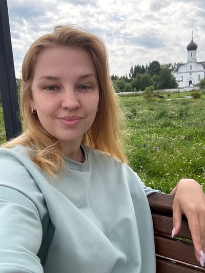 In Breytovo, Russia, a young woman with blonde hair and freckles is taking a selfie while sitting on a wooden bench. She is wearing a light blue sweatshirt. Behind her, a white church with a dark dome stands amidst a field of green grass dotted with small white and pink flowers. Trees and a cloudy sky complete the scene.