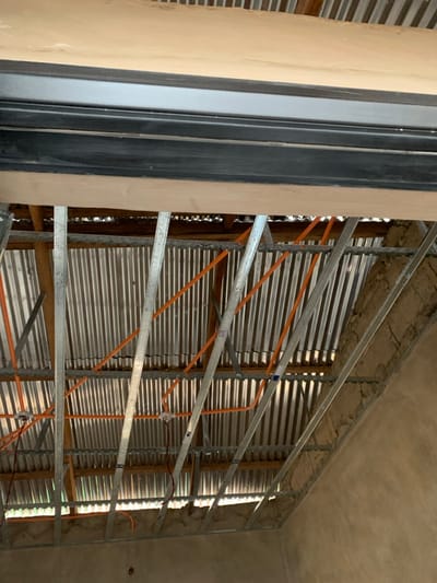 The image displays an interior view of an unfinished building in Dekpor Horme, Ghana, focusing on the ceiling and roof structure. The upper portion of the frame features a light-colored wooden panel, possibly plywood, with a black metallic trim or frame running horizontally beneath it. Below this, a grid of silver-colored galvanized metal studs or furring channels forms the framework for a suspended ceiling. Integrated within this metal framework are multiple runs of bright orange electrical conduits, routed across the scene. Several white electrical junction boxes are visible, from which dark electrical wires extend downwards. Above the metal framework, the corrugated galvanized iron roof is clearly visible, with visible wooden purlins or support beams underneath the corrugated sheets. To the right of the frame, an unpainted or unfinished light brown wall, appearing to be concrete or stucco, rises towards the roofline. No individuals are present, and no active construction work is depicted. The scene is illuminated by ambient daylight, reflecting off the corrugated roof.