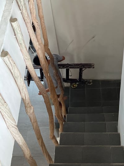 The image shows an interior in Singakerta, Indonesia, with a person reflected in a mirror. The person is wearing a gray long-sleeved shirt, and only their legs, torso, and part of their head are visible in the reflection. Behind the person and their reflection, there's a dark wooden chair with a small floral arrangement on the upper part of the back. The backdrop includes a light-colored wall to the left and a dark tiled wall to the right. In the foreground are several light-colored, rough-textured wooden branches laid out horizontally on a white surface.