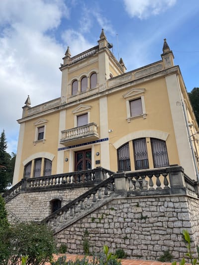 The image depicts the Centro Musical San Rafael, located in the city of Buñol, Spain. The building is a multi-story structure with a pale yellow facade and white architectural trim, set against a partially cloudy blue sky. It features an eclectic or neoclassical architectural style with decorative elements.
The main entrance is positioned centrally on the second level, accessible via a broad, symmetrical stone staircase with balustrades. The staircase is built on a rough-hewn stone retaining wall. Above the entrance, a blue sign with white lettering reads "CENTRO MUSICAL SAN RAFAEL." Directly above this sign and the entrance, a small balcony with a white balustrade protrudes from the facade. The entrance itself consists of ornate dark wood double doors.
Windows on the lower and upper levels vary in shape, including rectangular and arched designs. Most windows are fitted with closed internal blinds and some, particularly on the lower level, are protected by decorative black iron grilles. The building features white pilasters and cornices, and a prominent central tower-like projection above the main section of the roofline. This projection contains three arched windows with closed blinds. The roofline is adorned with multiple pointed finials or pinnacles, and a small satellite dish and an antenna mast are visible on the roof.
To the left of the building, a mature pine tree is visible, extending above the roofline. In the foreground, at the base of the stone staircase and retaining wall, there are landscaped garden beds with green bushes and low-growing vegetation. A terracotta-colored tiled pathway or ground surface is visible at the very bottom of the frame. No individuals are present in the scene. Two small white security cameras are mounted on the facade near the main entrance.