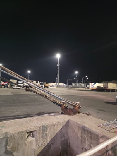 This is a night-time, long-shot, ground-level photograph taken at a port in Ravenna, Italy. The foreground is dominated by a thick, worn concrete structure, possibly a quay or dock, with a recessed area containing a metal ring and various cables. Several heavy, frayed ropes are attached to a rusted metal bollard on top of the concrete structure, extending diagonally upwards and out of the frame. The middle ground shows a paved port area illuminated by bright floodlights mounted on tall metal towers, creating stark contrasts and long shadows. Several large vehicles are present, including trucks with shipping containers, a white passenger vehicle, a tractor, and a yellow excavator. In the background, more floodlights and some industrial buildings or storage structures are visible, along with a dark night sky. The scene depicts an active, albeit currently unpopulated, port facility.