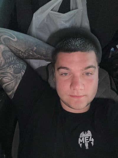 The image shows a close-up selfie of a young man with short, spiky dark hair and light blue eyes. He is wearing a black t-shirt with a white logo on the left chest area that appears to be stylized text and a winged figure. The man's left arm, adorned with a large, detailed tattoo, is resting behind his head. The background is dark and out of focus, with some indistinct shapes that could be furniture or other objects. There is a white plastic bag visible in the upper portion of the image. The lighting is dim, creating shadows. The image does not contain any recognizable landmarks or features that would allow for the identification of its location within the city of Husiatyn, Ukraine.
