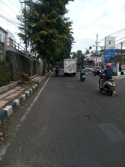 The scene depicts a street in the city of Khajuripada, India, under an overcast sky. The asphalt road, showing patches and white lane markings, occupies the central and right portions of the frame. To the left, a concrete sidewalk with black and white checkered curbing borders a stone retaining wall. A line of mature trees with green foliage stands along the left side, behind the sidewalk and wall.
Multiple vehicles are in motion, traveling away from the camera. In the foreground right, an individual wearing a blue shirt, shorts, and a pink helmet operates a light-colored scooter with a visible license plate number "4279." To the left and slightly ahead, another person on a dark scooter, wearing a grey helmet and dark attire, proceeds. Further into the scene, a white box truck with a red bumper and partially obscured rear license plate (beginning with 'B') is positioned in the left lane. Behind the truck, two additional individuals on dark motorcycles are visible, one wearing a red jacket.
The background on the right side reveals a commercial urban environment featuring multiple buildings, utility poles, overhead power lines, and a cellular tower. Prominent signage includes a large white billboard displaying text such as "PESONA," "METRO," "15.000," "34 - 36," and "DO NOT LOAD." A red circular 'no entry' sign is also visible. A traffic light assembly is present above the distant intersection.