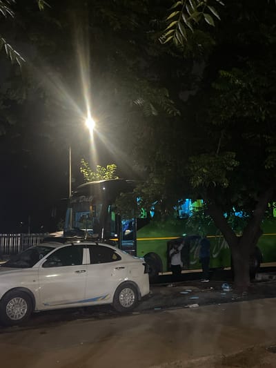 In Kajheri, India, at night, a white sedan is parked on the side of a street. A large green and black bus is parked behind the sedan. The bus has writing on its side that appears to say "Kam Dali". A street light shines brightly above and to the left of the bus, casting rays and lens flares. Trees with dark leaves overhang the scene. Two people are visible near the entrance of the bus. One person in a white shirt and dark pants is standing by the bus door, and another person in a blue shirt and dark pants is standing near them. The ground is paved with some debris and white markings.