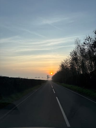 This image displays a rural road scene near Baschurch, United Kingdom, at either sunrise or sunset. A dark asphalt road, marked with a dashed white centerline, extends from the immediate foreground into the distant horizon. A prominent, intense orange sun is centered on the horizon, positioned slightly to the right of the road's vanishing point. The sky transitions from a pale blue in the upper portion, featuring faint, wispy clouds, to a warm orange and yellow gradient near the horizon. To the left of the road, a dark green, dense hedge runs parallel, with a layer of ground mist or fog obscuring the bases of distant silhouetted trees beyond it. To the right of the road, a thick line of bare deciduous trees and shrubs with dark branches forms a dense barrier against the skyline. No human figures, vehicles (other than the implied camera platform), or animals are present.