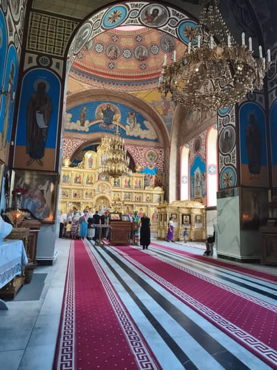 The image depicts the interior of a richly decorated Eastern Orthodox church in Ocnița, Moldova. The scene is dominated by ornate golden iconostases, colorful frescoes on the walls and ceiling, and a large, elaborate chandelier. Several individuals, dressed in everyday clothing, are present within the church, some standing in groups, suggesting a religious service or a visit. A red carpet with a decorative border runs down the center aisle, leading towards the altar area. The architectural style and religious iconography clearly identify the location as a place of worship within the Orthodox tradition.