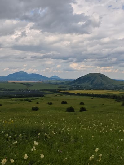 The image depicts a panoramic landscape featuring a verdant rolling field in the foreground dotted with small white wildflowers and green shrubs. In the mid-ground, the landscape transitions to more cultivated fields, including areas of yellow crops and a dark green treeline. Further in the distance, several distinct mountains rise against a sky filled with dramatic, overcast clouds. The largest mountain on the right is a rounded, cone-shaped peak. To the left, a range of mountains forms a jagged silhouette, with a city visible in the valley between them and the mid-ground. The overall impression is of a natural landscape with human settlement present in the distance, under a dynamic sky. The location is identified as Etoka, Russia.