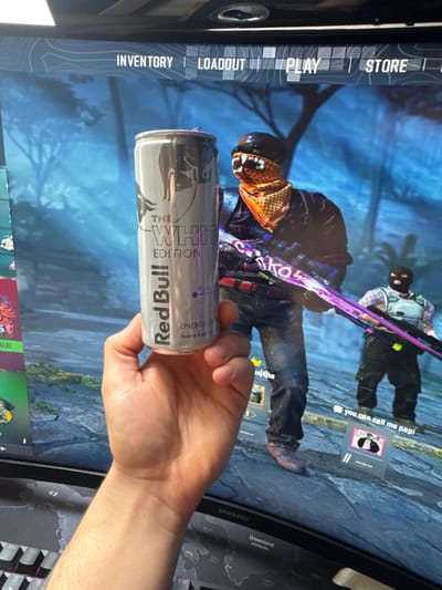 The image depicts an indoor scene in Pfaffenhofen an der Ilm, Germany, centered on a human right hand holding a silver Red Bull "The White Edition" energy drink can. The can is open, with the pull tab elevated, and features text including "Red Bull," "THE WHITE EDITION," "ENERGY DRINK," "Kokusnuss" (Coconut), and "Belebt Geist und Körper" (Revitalizes Mind and Body). In the background, a computer monitor displays a video game featuring two armed characters in what appears to be a forest or outdoor environment. The most prominent game character wears a dark cap, an orange and black patterned bandana covering the lower face, a dark jacket, and jeans, holding a long firearm with a distinct purple, pink, and white design. A second, partially obscured game character is visible further back, wearing a dark balaclava and a light-colored tactical vest, also appearing to hold a firearm. The monitor's top navigation bar displays "INVENTORY | LOADOUT | PLAY | STORE." A "SAMSUNG" logo is visible on the bottom bezel of the monitor. A black mechanical keyboard, featuring a key labeled "Greenland," is partially visible at the bottom of the frame.