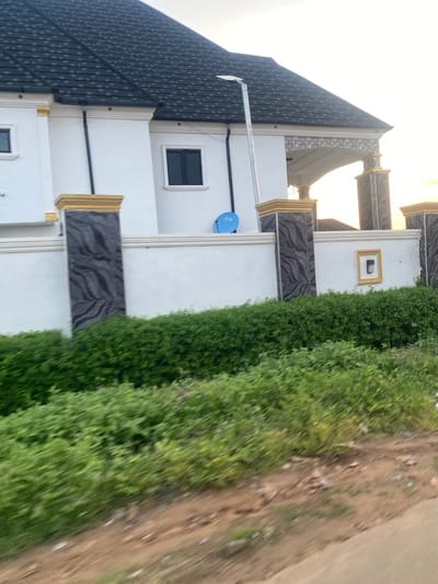 The image captures a static scene of a property in Enwan, Nigeria, featuring a white building with a dark roof, partially obscured by a perimeter wall, viewed from a roadside perspective.
The building exhibits a multi-pitched roof composed of dark gray asphalt shingles. Its exterior walls are white with white decorative cornices. Two dark-paned rectangular windows are visible on the upper sections of the building. A black vertical drainpipe or conduit is affixed to the left portion of the facade. A small, round, blue satellite dish is mounted on the white wall below one of the windows. A slender silver pole, likely a streetlight, stands upright in front of the building.
A white perimeter wall stretches horizontally across the mid-ground. This wall is punctuated by decorative pillars that feature golden-yellow caps and bases, framing dark gray textured panels with undulating patterns. Towards the right, a portion of an arched entrance or portico, supported by similar pillars and featuring geometric patterns on its soffit, is visible. A small, rectangular niche with a golden-yellow frame and an embedded light fixture is present on the far right section of the white wall.
In the foreground, a dense band of vibrant green foliage, comprising bushes and plants, lines the base of the perimeter wall. Below the foliage, an uneven strip of reddish-brown dirt and soil, containing scattered small debris, extends. A section of a dark, paved surface, likely a road, is visible in the bottom right corner. The sky is bright, with a soft orange-yellow gradient visible on the right horizon, suggesting either sunrise or sunset. No individuals or specific actions are depicted within the frame.