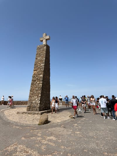 The image depicts a sunny day at Cabo da Roca, the westernmost point of mainland Europe, located in the municipality of Sintra, near Colares, Portugal. A prominent stone monument, a large cross atop a tapering stone pillar, stands in the center-left of the frame against a clear blue sky. The monument is surrounded by a paved area with intricate stonework patterns. A crowd of tourists is present, walking around the monument and enjoying the view. The people are dressed in casual summer clothing, with many wearing shorts, t-shirts, and hats. Some are taking photographs, while others are simply strolling. The environment is coastal, with a hint of the ocean visible in the background, beyond a low stone wall. The overall atmosphere is one of a popular tourist attraction on a bright, clear day.