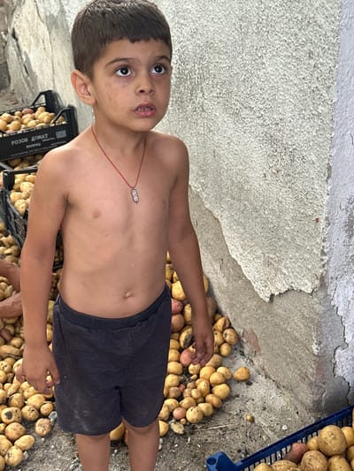 A young boy stands in front of a textured, light-colored wall in Mlekarevo, Bulgaria, looking upwards and to the right with a slightly open mouth. He is shirtless, wearing dark shorts, and a red string with a rectangular pendant around his neck. He is surrounded by potatoes. To his left, a black plastic crate is filled with potatoes, with text in a foreign language on its side. More potatoes are scattered on the ground around him, with a blue plastic crate visible in the lower right corner, also containing potatoes. The ground appears to be gravel and dirt. The lighting suggests it is daytime.