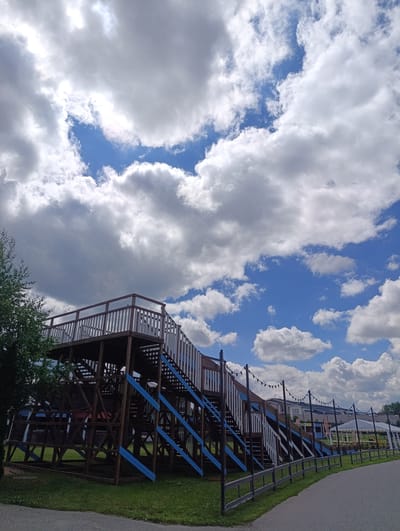 In Ivanovskoe, Russia, a large wooden structure with stairs and platforms, likely a playground or event space, is visible under a sky filled with dramatic clouds and patches of blue. The structure features white railings and blue accents on its supports. A paved walkway runs alongside a grassy area in front of the structure, with a fence separating them. In the background, there are buildings, a white tent, and string lights, suggesting a public outdoor area. A tree stands to the left of the wooden structure.