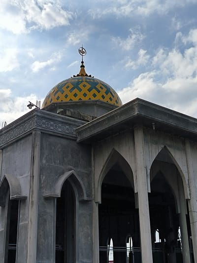 The image shows a partly constructed mosque in Blangbitra, Indonesia. The building features a dome covered in a geometric pattern of yellow and turquoise. The facade is made of grey concrete with arched entrances supported by pillars. The building appears to be under construction, with the interior still exposed. The sky is visible in the background, dotted with white clouds.