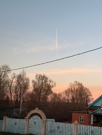 The image shows a landscape in Staraya Yum'ya, Russia. The sky transitions from light blue to a pale orange-pink. A contrail is visible in the sky, bisected by a black wire running diagonally across the frame. Below the sky, there's a row of trees with bare branches silhouetted against the sky. A red house with a green roof is partially visible on the right. A light blue, wooden fence with an arched top is in the foreground on the right.