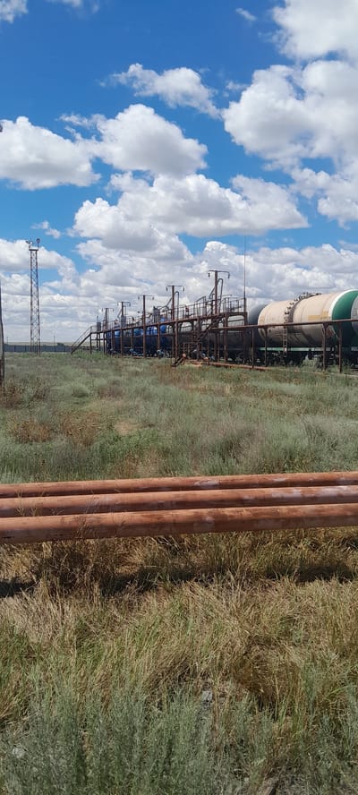 This is a medium shot of an industrial area with oil or gas transport infrastructure, located in the city of Embi, Kazakhstan. The scene is dominated by large, rust-colored pipes in the foreground, lying horizontally in the dry, grassy terrain. In the background, a network of elevated metal structures, likely part of a loading or unloading facility, supports multiple tanker cars. These cars appear to be either blue or white and green. A tall, metal tower with a lighting fixture is visible to the left, adding to the industrial feel. The sky above is a bright blue, dotted with fluffy white clouds. The overall impression is one of arid, industrial landscape under a partly cloudy sky. There are no people visible in the image.