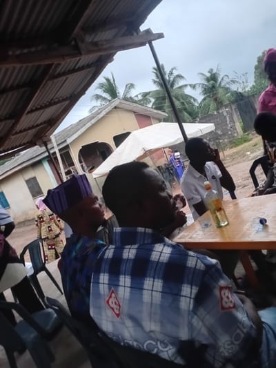 The image depicts an outdoor gathering in Ipota, Nigeria, possibly at a local market or community event. The scene is partially covered by a corrugated metal roof structure supported by wooden beams. In the foreground, two men are seated, with the focus on the man in the lower right, wearing a blue and white plaid shirt with a red emblem. To his left, another man, mostly obscured, appears to be speaking. A table is visible, with a bottle of clear liquid, a glass, and a plastic water bottle on it.
In the background, a white canopy tent is set up, suggesting a stall or gathering point. Several people are visible around and behind the tent. One person wears a brightly patterned traditional African outfit and a purple cap. Palm trees are visible in the distance, along with a brick wall and parts of buildings, indicating a rural or semi-urban environment. The sky is overcast. The overall impression is of a casual community gathering.