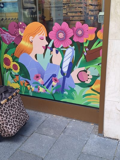 The image depicts a shop window display featuring a colorful illustration of a woman applying makeup amidst a floral and foliage background. The woman has orange hair and is holding a lipstick and a compact. The background consists of large, stylized pink and purple flowers, green leaves, and sunflowers. To the left of the window, a leopard-print bag with wooden handles is visible. The display is positioned in front of a shop interior filled with shelves of products, suggesting it might be a pharmacy or cosmetic store. The exterior pavement in front of the window is made of gray stone tiles. Several cigarette butts are scattered on the pavement near the base of the window. The architecture visible in the background of the shop interior suggests a European setting. Based on the provided context, this scene is located in Bochum, Germany.