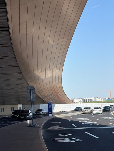 The image depicts a street scene in Shunyi District, China. A curved, tiled building facade occupies the left side of the frame, contrasting with the clear blue sky above. Adjacent to the building is a multi-lane road with traffic, including cars and a van. Road markings are visible on the asphalt surface. A construction crane appears in the distance. A street sign and pole are situated near the curb, with Chinese characters visible on the sign.