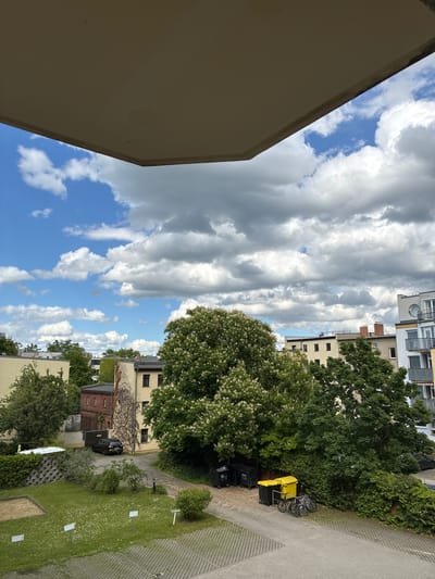 The image presents an outdoor scene, likely taken from an elevated position such as a balcony, in a residential area of Cottbus-Chóśebuz, Germany. On the left side of the frame, a portion of a light-colored building structure is visible. The sky is partly cloudy with blue patches.

The central and right parts of the image show a streetscape with a mix of buildings and greenery. Buildings of varying heights and architectural styles are present, some with brickwork or ivy climbing their facades. Trees and bushes are interspersed among the buildings. On the right is a paved area, a strip of grassy lawn, and what appear to be parked cars, bins, and bicycles. There is no evidence of people visible in the scene.