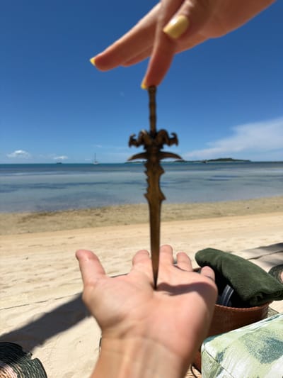 In Baan Thong Krut, Thailand, a person's hands are holding a miniature sword over a sandy beach. The sword is brass-colored and has a decorative hilt with ram-like horns. The right hand, with yellow-painted fingernails, is positioned above the sword, with two fingers extended. The left hand, palm up, is positioned below the sword, appearing to catch it. The background shows a calm, blue ocean with two sailboats in the distance, and a clear, bright blue sky. Part of a woven basket containing a green cloth and other objects, and a patterned fabric item, are visible on the right side of the frame, suggesting the scene is set near a lounging area.