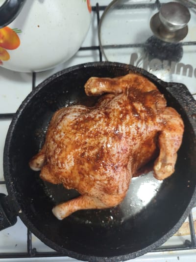 The image shows a whole, raw chicken seasoned with spices and placed in a black cast-iron skillet. The chicken is positioned in the center of the skillet, filling most of its surface. Its skin is a pale beige with patches of red and brown seasoning. The legs and wings are tucked in. The skillet is on a white stovetop with black grates. To the left of the skillet, a portion of a white pot with a decorative floral pattern is visible. To the right of the skillet, a clear glass lid with a silver handle is placed on the stovetop, with a metallic surface reflected on it. The overall setting appears to be a kitchen. No people are visible, and the activity depicted is food preparation, specifically the seasoning of a chicken before cooking. The lighting suggests it could be daytime. There is no visible text in the image.