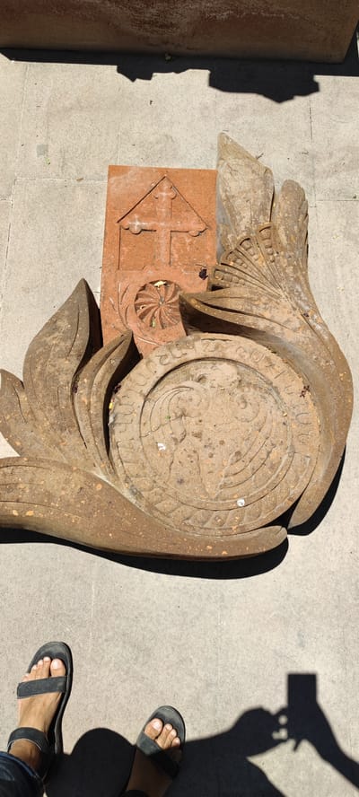 This media file shows a pair of feet wearing black sandals standing on a concrete surface. In the background, there is a weathered stone carving that appears to be part of a monument or sculpture. The carving features intricate details, including what looks like a cross on a triangular base, a circular motif with floral patterns, and decorative leaf-like elements. The stone itself is reddish-brown and shows signs of age and exposure to the elements. The lighting suggests it is daytime, with shadows cast on the ground, indicating a sunny environment. The ground is paved with light-colored concrete slabs. The scene is outdoors, likely in a historical or cultural site given the nature of the stone carving. There is no visible text in the image. The overall mood is serene and contemplative, focusing on the details of the ancient artwork and its setting.