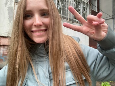 A young woman with long, blonde hair is taking a selfie outdoors. She is wearing a light blue hooded jacket. Her left hand is raised, forming a peace sign. She is smiling and looking at the camera. The background appears to be a building with barred windows, suggesting an urban environment. Based on the provided information, the location is Yuzhno-Sakhalinsk, Russia.