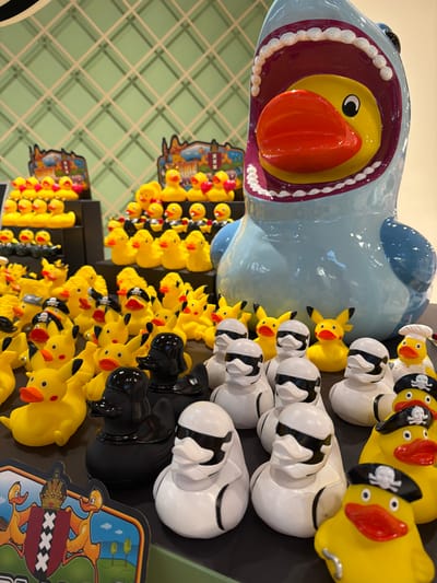 This media file features a variety of rubber ducks and Pikachu figurines displayed in a store setting. The main subject is a large, light blue shark-shaped object with an open mouth, revealing a yellow duck inside. Surrounding the shark are rows of rubber ducks, some yellow with pirate hats, and others white with black stripes. Numerous yellow Pikachu figurines are also visible, arranged in rows. In the background, a wall with a green grid pattern is partially visible. The objects appear to be for sale or display, likely in a novelty shop or toy store. The lighting suggests an indoor environment. There is no visible text in the main portion of the image, though two colorful signs with cartoon ducks and what appears to be a coat of arms with "XXX" are present at the bottom right. The overall scene evokes a playful and whimsical atmosphere.