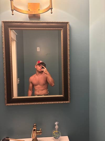 The image shows a shirtless man in a bathroom, taking a mirror selfie. He is wearing a baseball cap and a gold chain. The bathroom has teal walls, a gold-framed mirror, and a gold faucet. There is also a bottle of hand soap visible on the counter. The location is identified as Panama City Beach, United States of America.