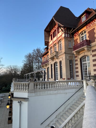 The image presents a building's exterior with distinct architectural styles. One section showcases a half-timbered design with red and white accents under a dark roof, while the adjacent part displays classic stone facades with symmetrical windows. A white balustrade runs along the side of a patio or walkway. Bare trees suggest a cool season. A clear blue sky is visible in the background.