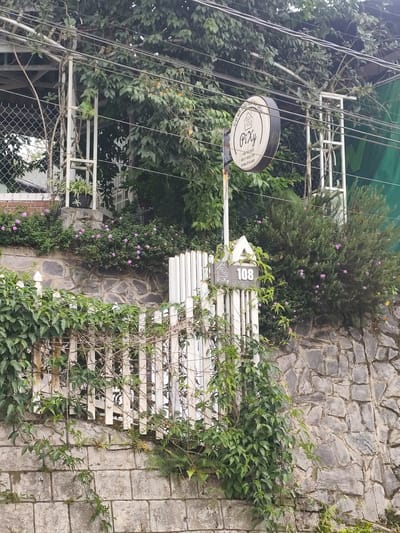 The image shows a stone wall topped with a white picket fence overgrown with greenery. Beyond the fence, there's a sign that reads "Pixy" with phone numbers below it. Further back, lush green trees and foliage dominate the scene, with some electrical wires strung overhead. A white structure, possibly part of a building or canopy, is visible through the trees. A white pole with a triangular top displays the number "108". The setting appears to be a natural, perhaps slightly overgrown, outdoor area. The details suggest a location in or around Lang Biang - Đà Lạt, Vietnam, given the presence of lush vegetation and the architectural style.